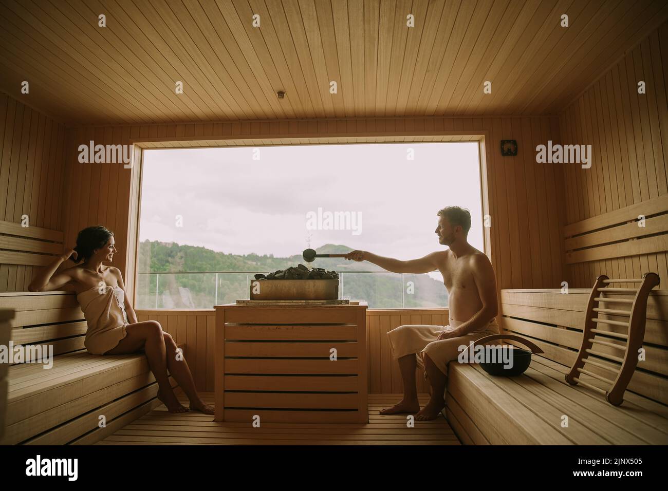 Handsome young couple relaxing in the sauna, while man pouring water on hot rocks Stock Photo