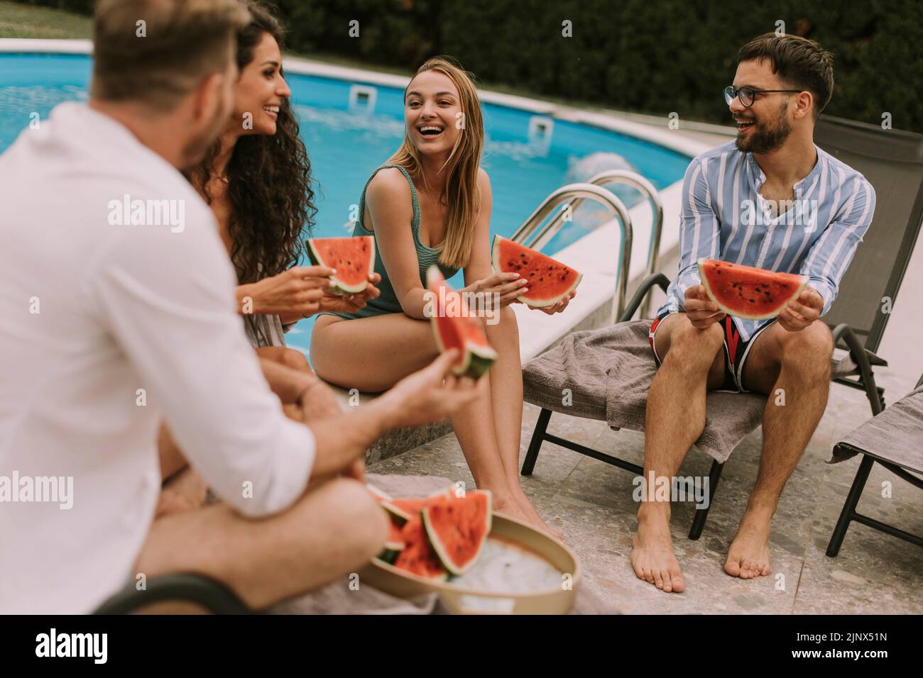 Group of young people sitting by the swimming pool and eating watermelon in the house backyard Stock Photo