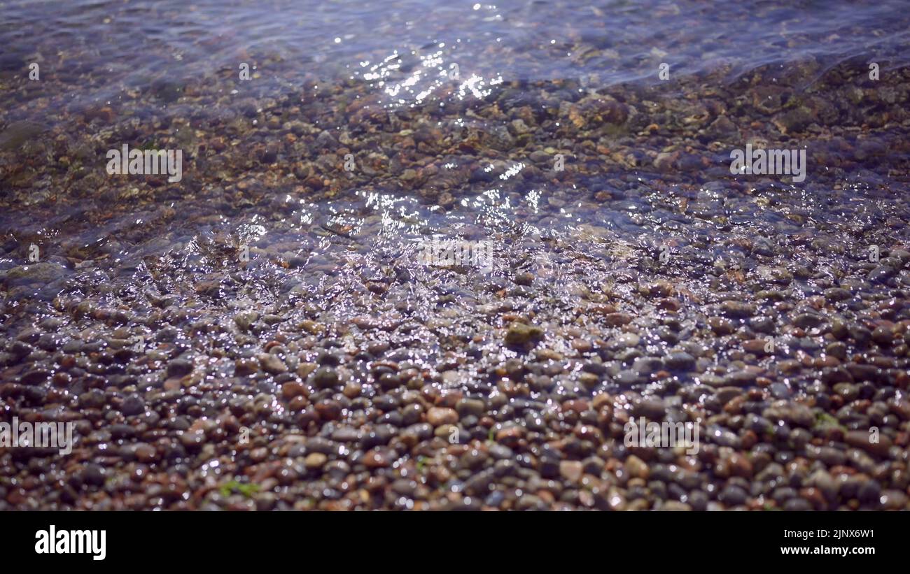 Close-up of pebble beach with transparent waves with sun glare on the water. Close up of stony sea shore. Natual background of a calm sea in the surf Stock Photo