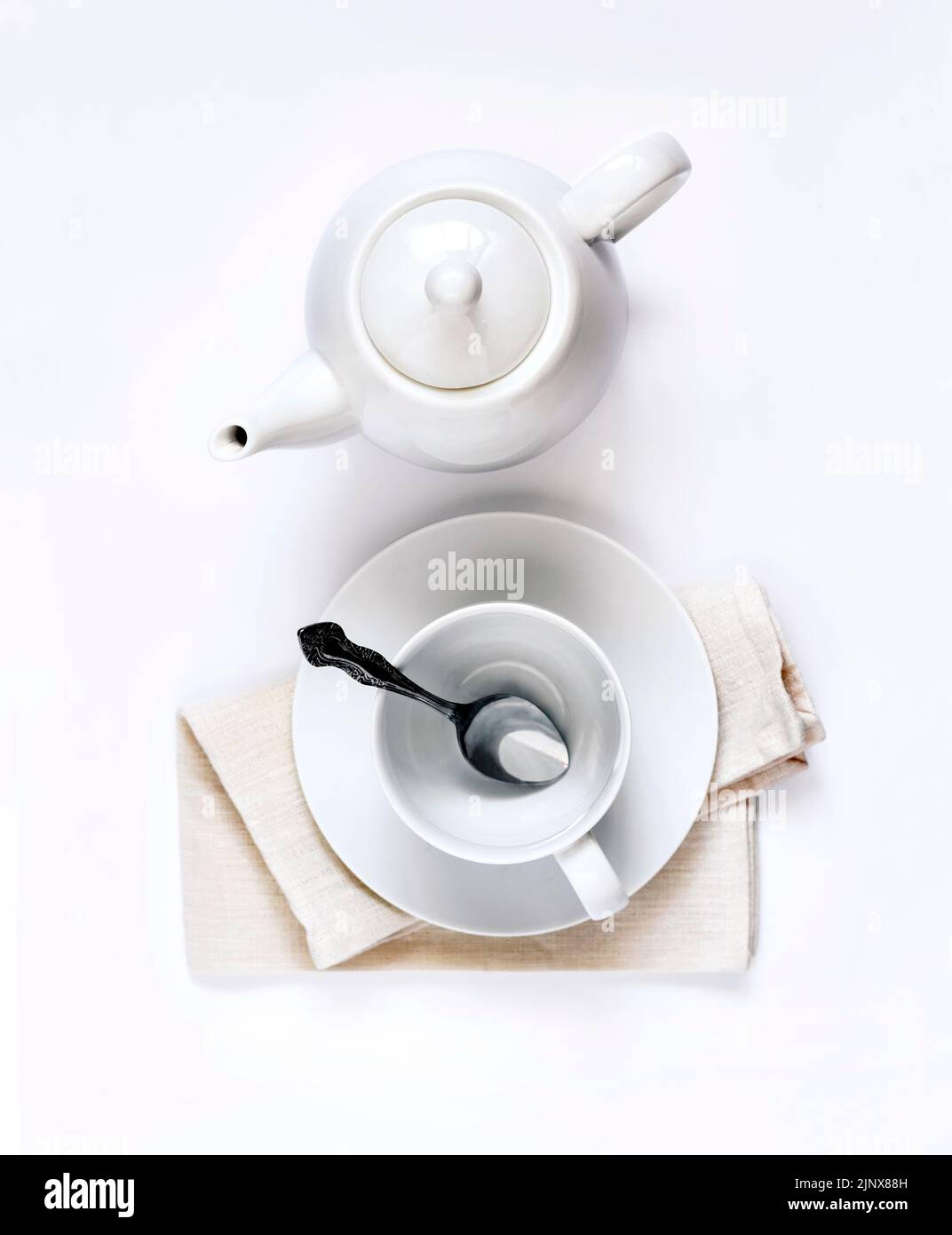 White teapot and empty teacup with spoon on pale beige cloth isolated on white background. Flat lay, top view Stock Photo