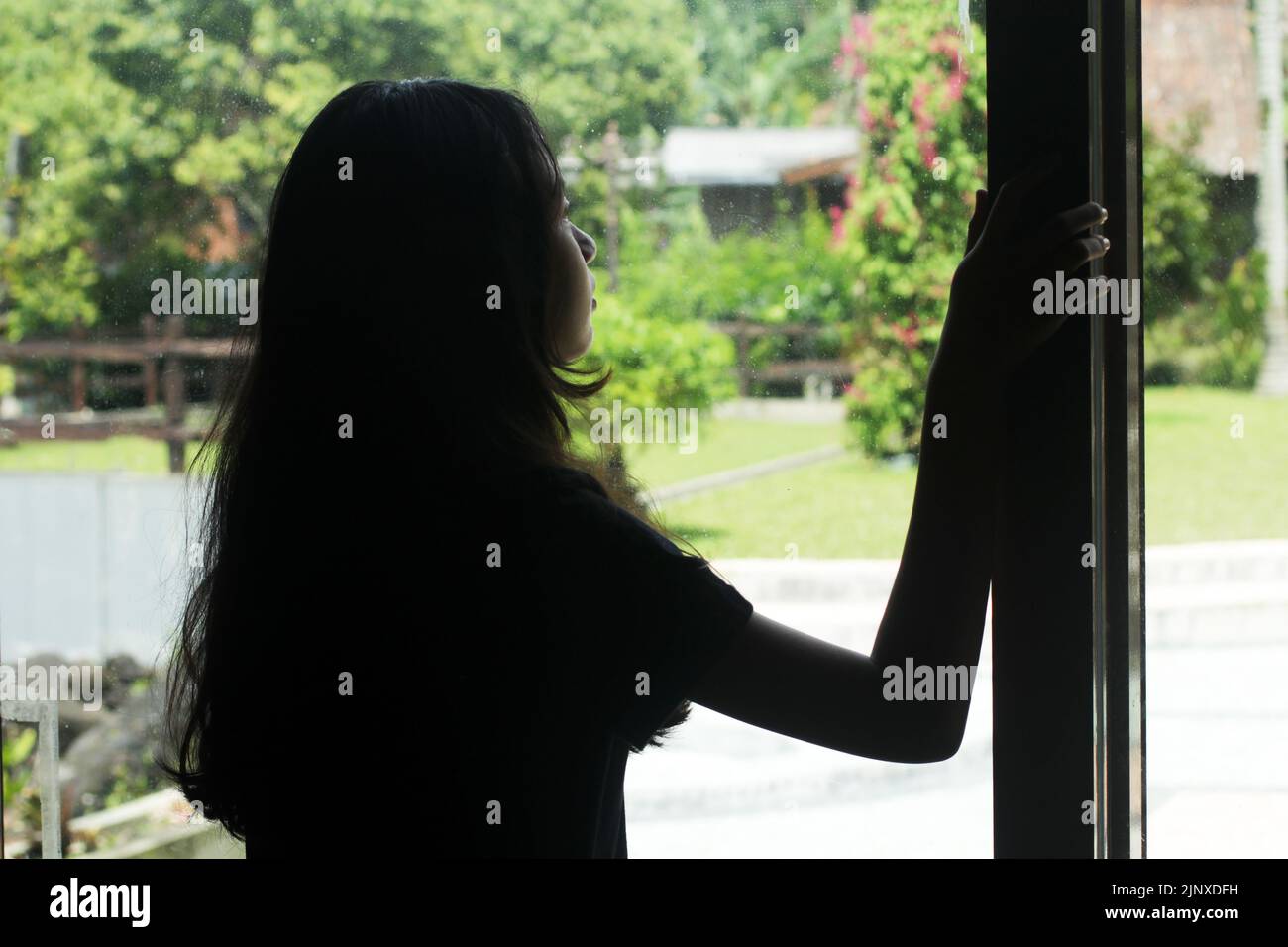 Silhouette of a girl with long hair, standing alone from behind, looking at the window day-dreaming thinking about life, deep in thought. Stock Photo