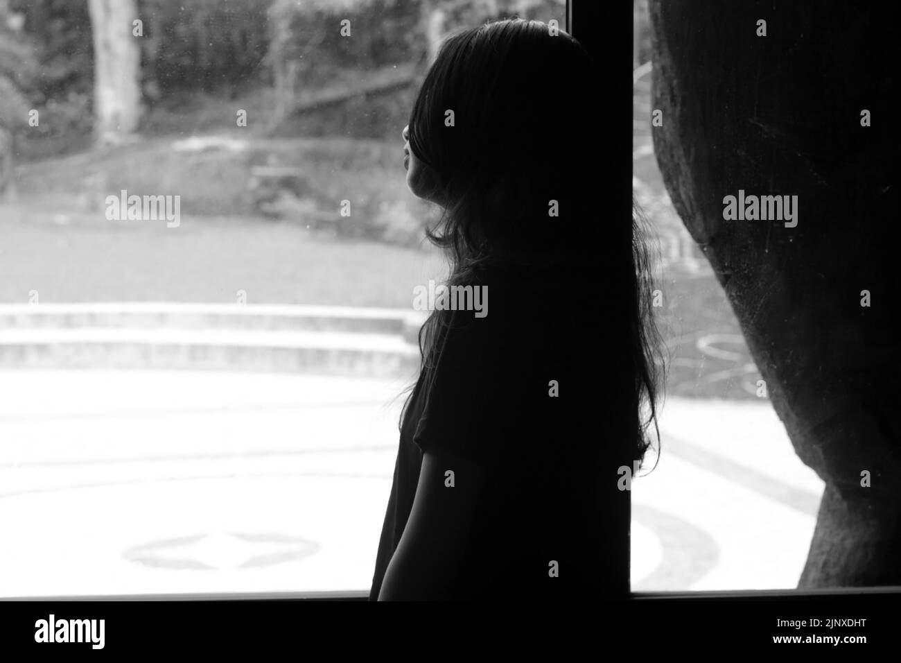 Silhouette of a girl with long hair, standing alone looking at the window day-dreaming thinking about life, deep in thought. In black and white. Stock Photo