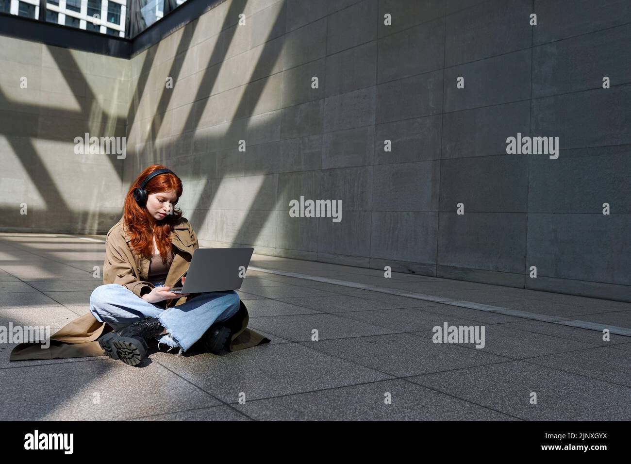 Teen redhead girl wearing headphones using laptop in city urban location. Stock Photo