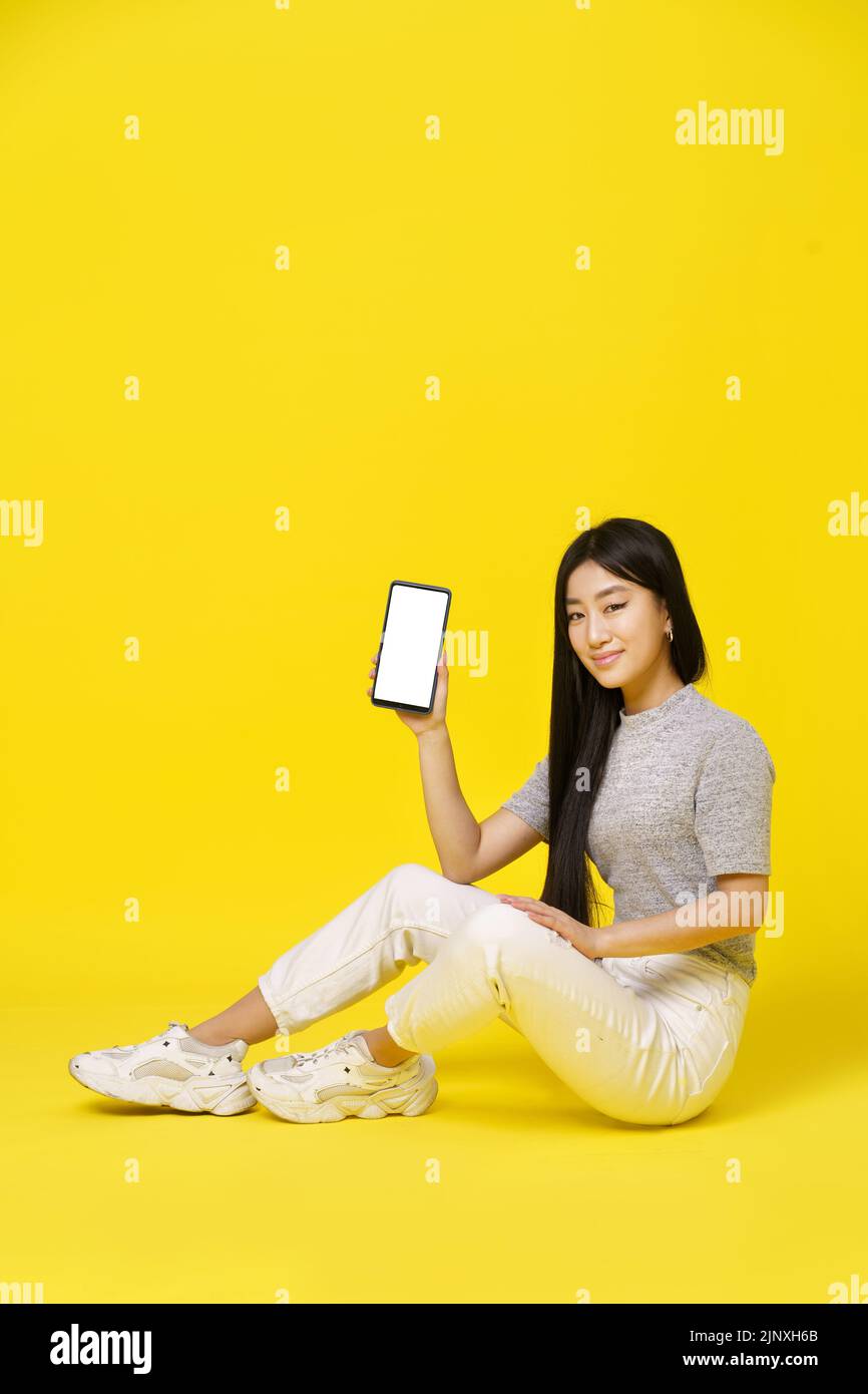 Charming asian young girl holding smartphone sitting on the floor showing a black screen isolated on yellow background. Great offer. Product placement. Mobile App Advertisement. Copy space.  Stock Photo