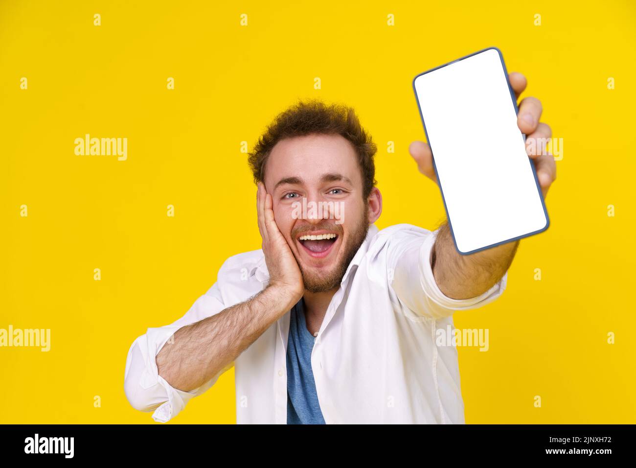 Great offer. Young happy man holding smartphone showing a white empty screen and exciting to win isolated over yellow background, celebrating success. Product placement. Mobile App Advertisement.  Stock Photo