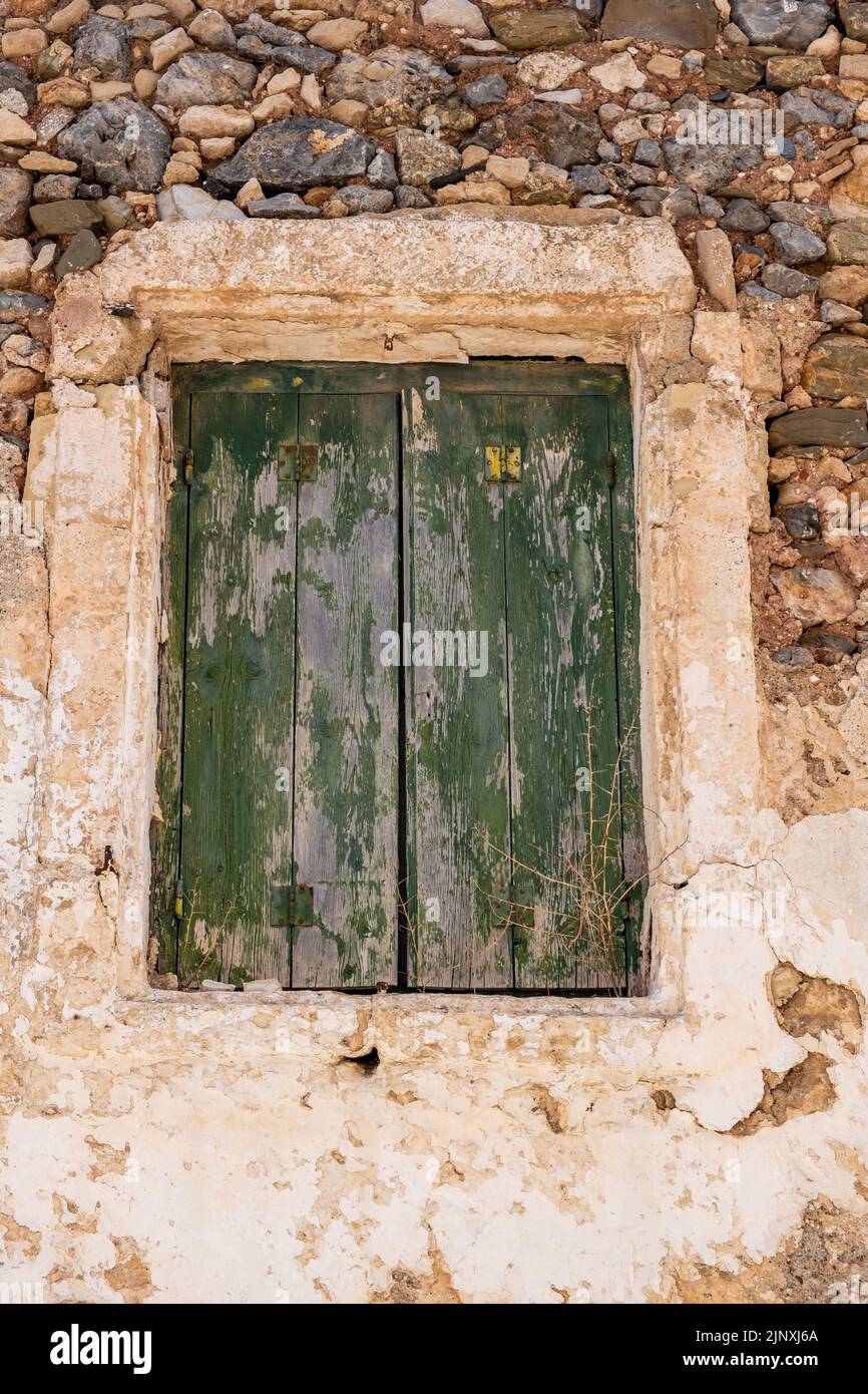 Aged wooden closed shutters, peeled stone wall Greek rural building facade. Abandoned house background, green painted window planks. Vertical Stock Photo