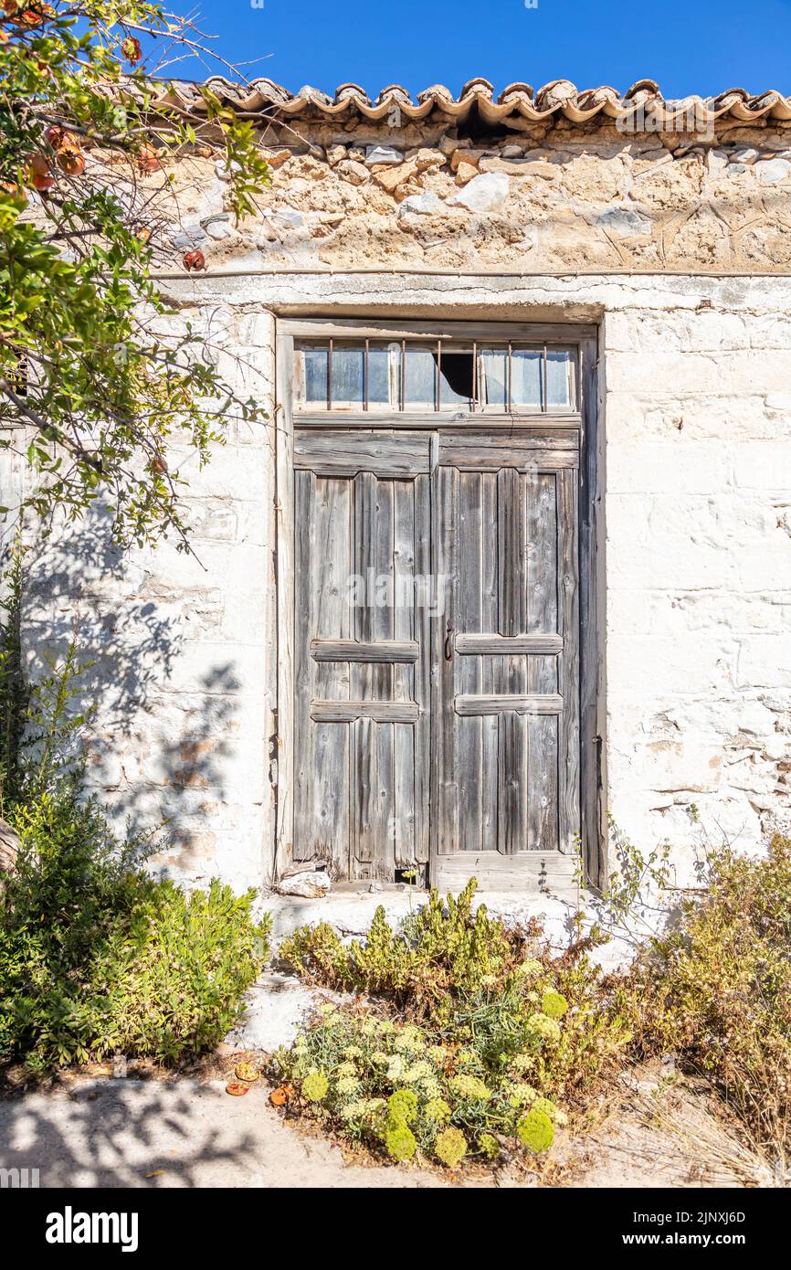 Old rural building wall facade, ceramic tiles on rooftop, wooden aged closed door with broken glass on top. Abandoned house, dry and green plants outd Stock Photo