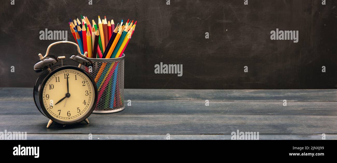 Time for school banner. Colorful pencils and alarm clock on classroom blue wooden desk, blackboard background, copy space Stock Photo