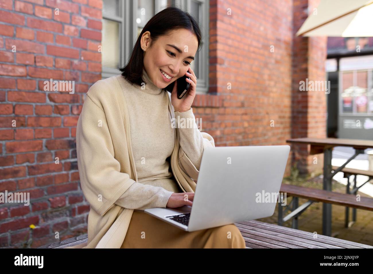 Smiling young Asian business woman talking on cellphone using laptop outdoors. Stock Photo