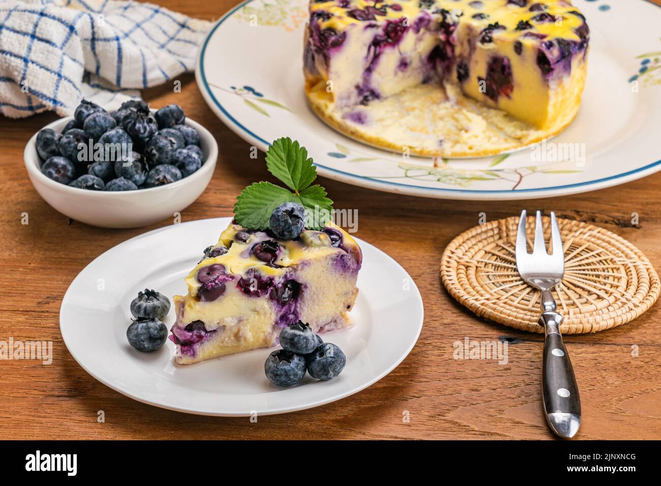 Blueberry yoghurt cake in white ceramic dish. Portion blueberry yogurt cake with blueberries in ceramic cup, metal fork on bamboo mat on wooden table. Stock Photo