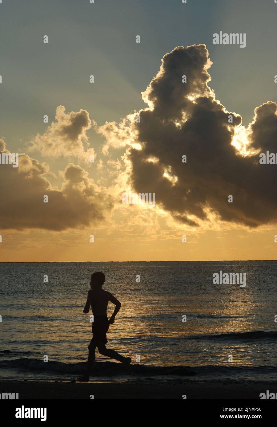 The silhouette of a young boy running along the seashore at dawn against a cloudy tropical sky Stock Photo