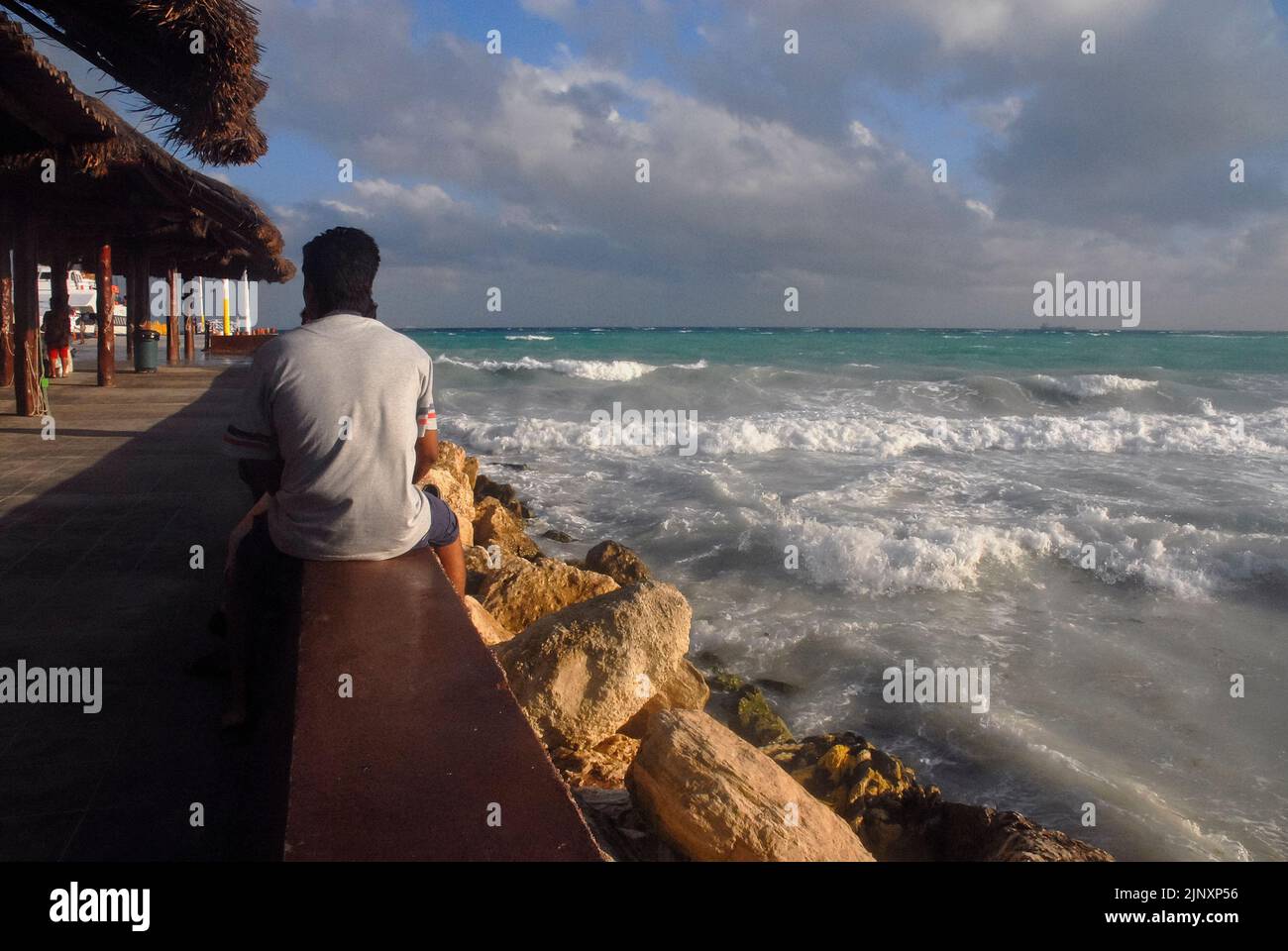 A rear view shot of a man sitting on a parapet as he watches the rough Caribbean Sea, and waves crashing on rocks in Mexico: Selective focus. Stock Photo