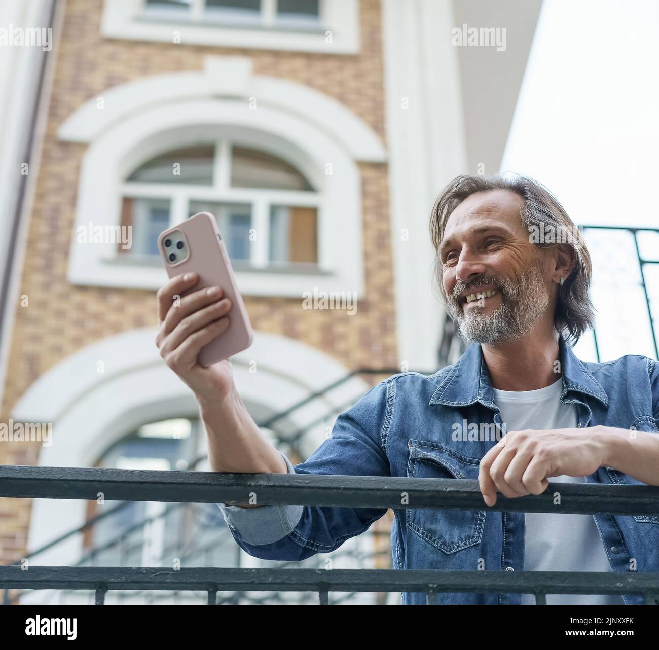 Happy middle aged grey bearded man laughing reading text message or funny social media post using smartphone standing in old town wearing casual. Travel concept.  Stock Photo