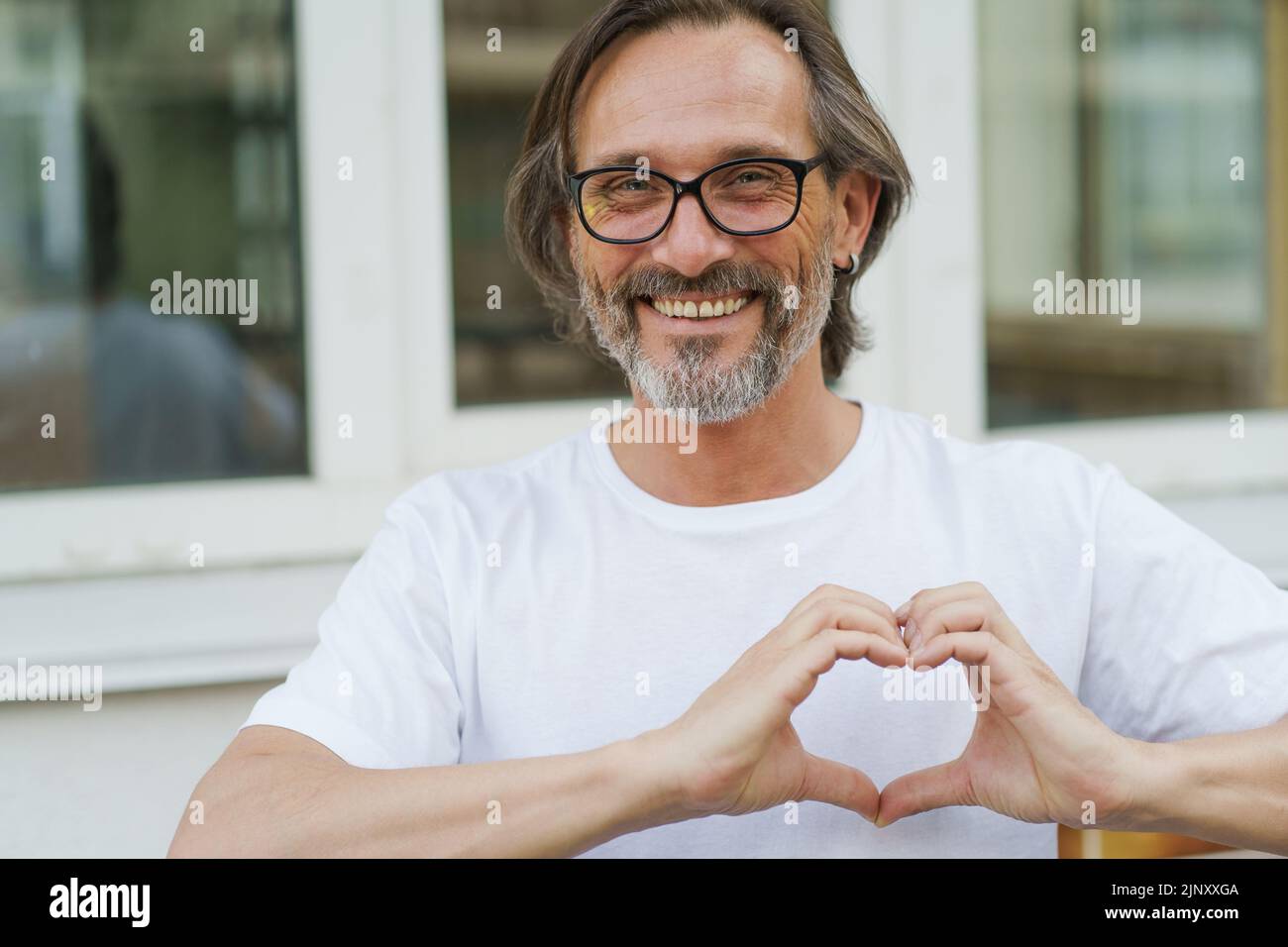 Gesturing heart, love, happy mature man in glasses smile standing outdoors wearing white t-shirt. Happy mature man in eye glasses and looking at camera sending love outdoor on a summer day. Stock Photo