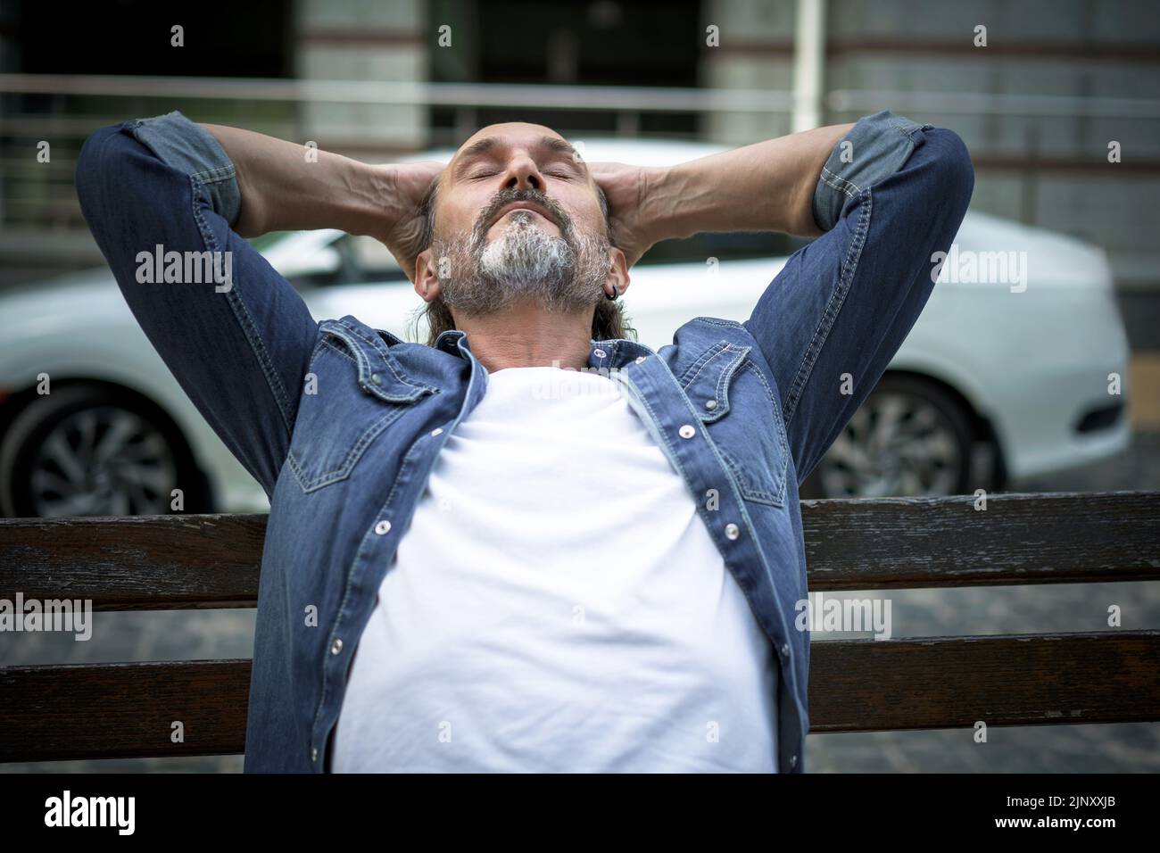 Relaxed, enjoying life handsome middle age man sits leaned back with hands behind his head on the bench outdoors at urban city background wearing denim shirt and white t-shirt. Travel concept.  Stock Photo