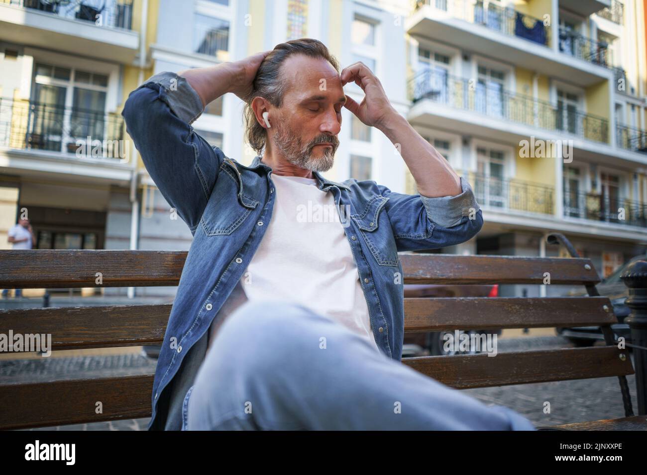 Handsome middle aged man sits on the bench listening music using wireless earphones, with hands lifted up fixing his long hair and eyes shut at old town background enjoying free time.  Stock Photo