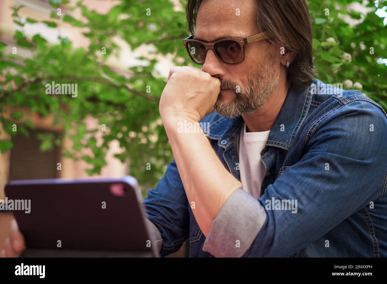 Puzzled up, sad middle aged man working outdoors using digital tablet. Handsome freelancer traveling man not happy checking work or social media sitting at city street cafe.  Stock Photo