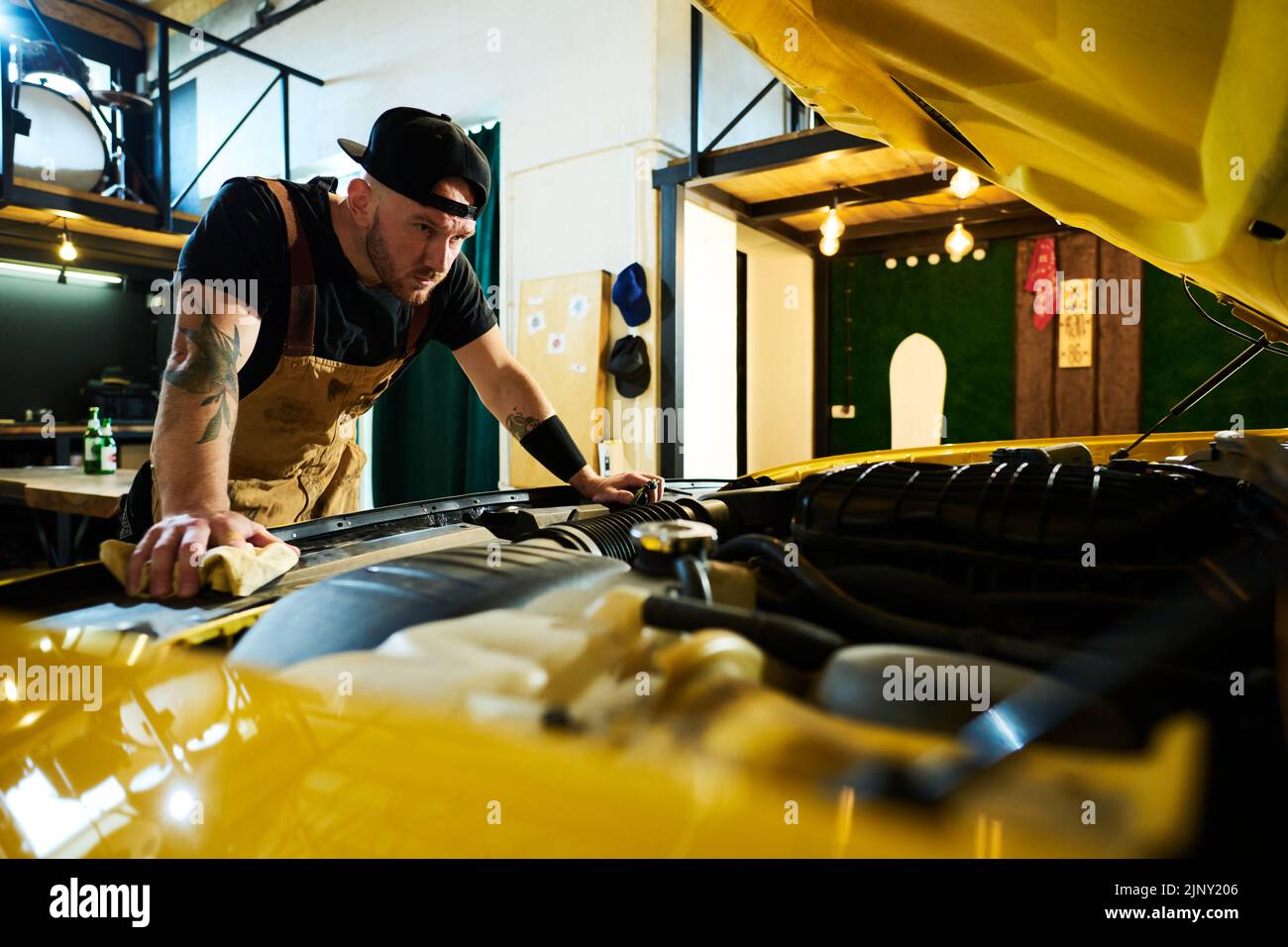 Young serious man in apron and baseball cap bending over open hood of car and carrying out checkup of engine in garage or workshop Stock Photo