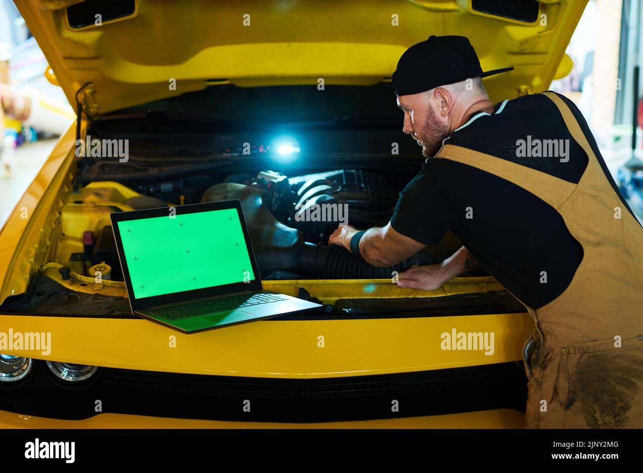 Young repairman keeping hand in car engine and looking at green screen of laptop while carrying