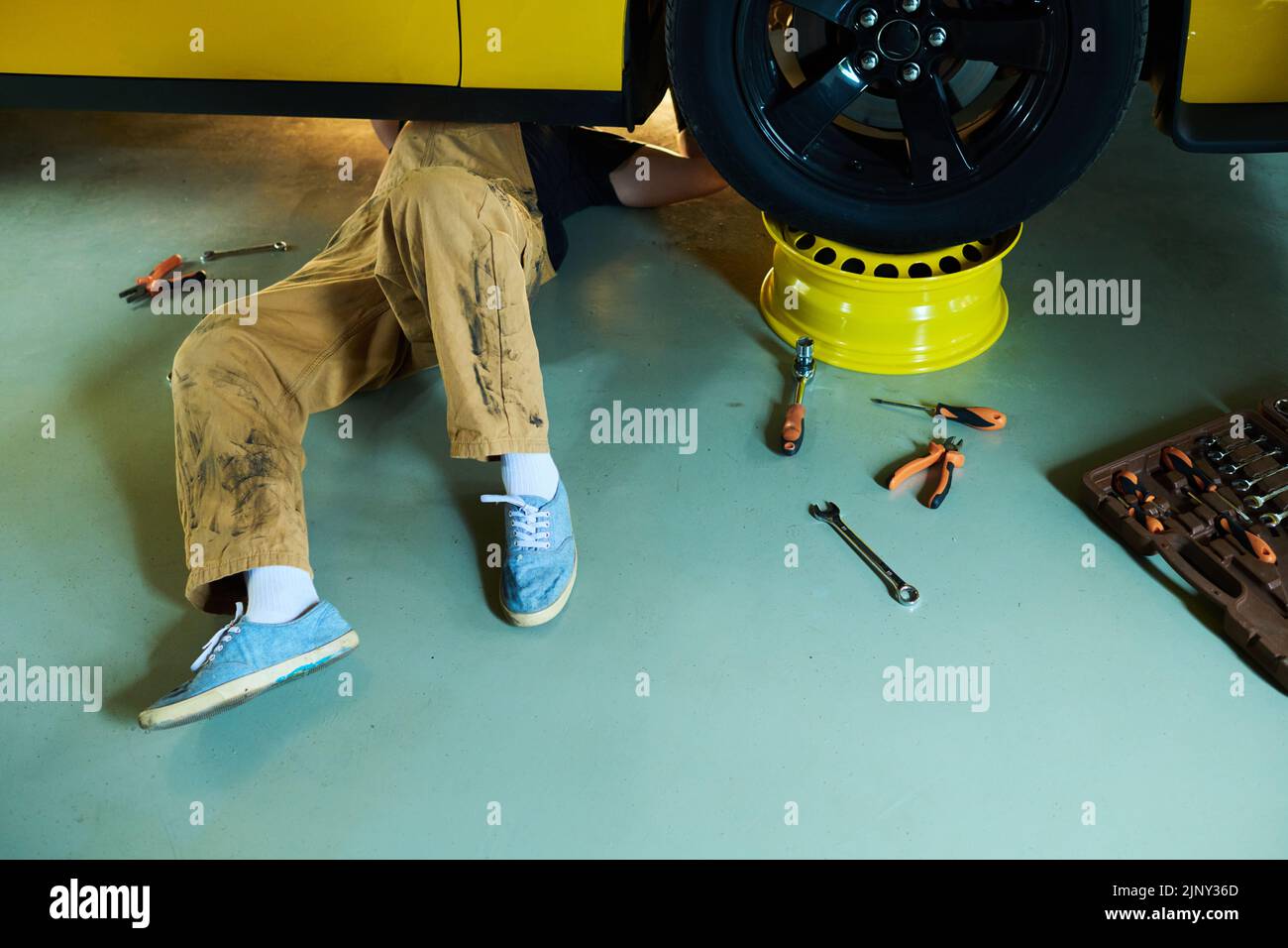 Legs of young repairman or technician in overalls lying under yellow electric car on the floor of workshop or garae and repairing it Stock Photo