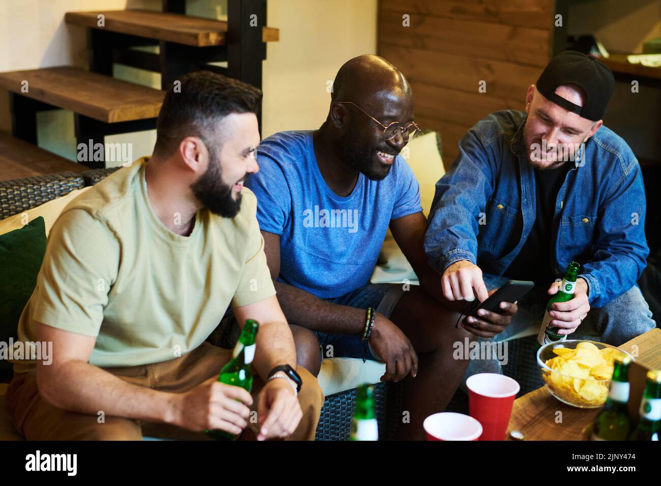 Cheerful buddies discussing photos in smartphone of African American guy while sitting by table with bottles of beer and snacks Stock Photo