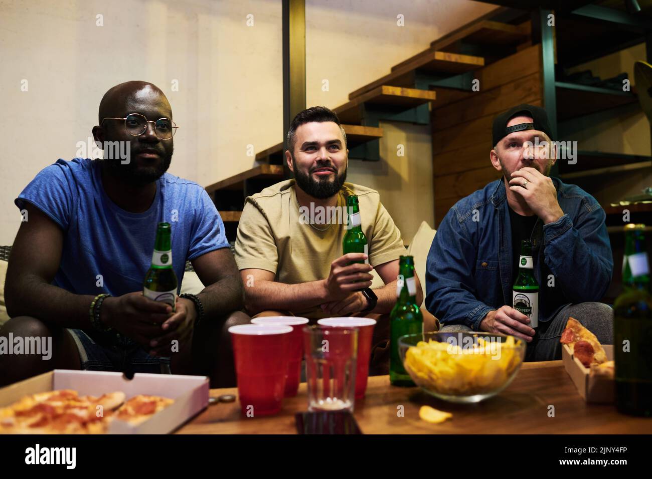 Three intercultural male football fans gathered by table with beer and snack in front of tv set in garage to watch broadcast of match Stock Photo