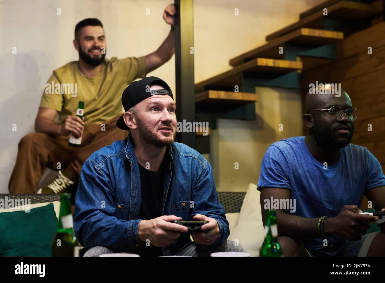 Young man in denim jacket and baseball cap and his African American buddy with joysticks playing computer game against guy with beer Stock Photo