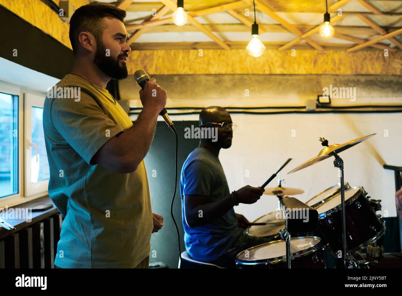 Young man with microphone by his mouth standing against black guy with drumsticks hitting cymbals and drums during repetition Stock Photo