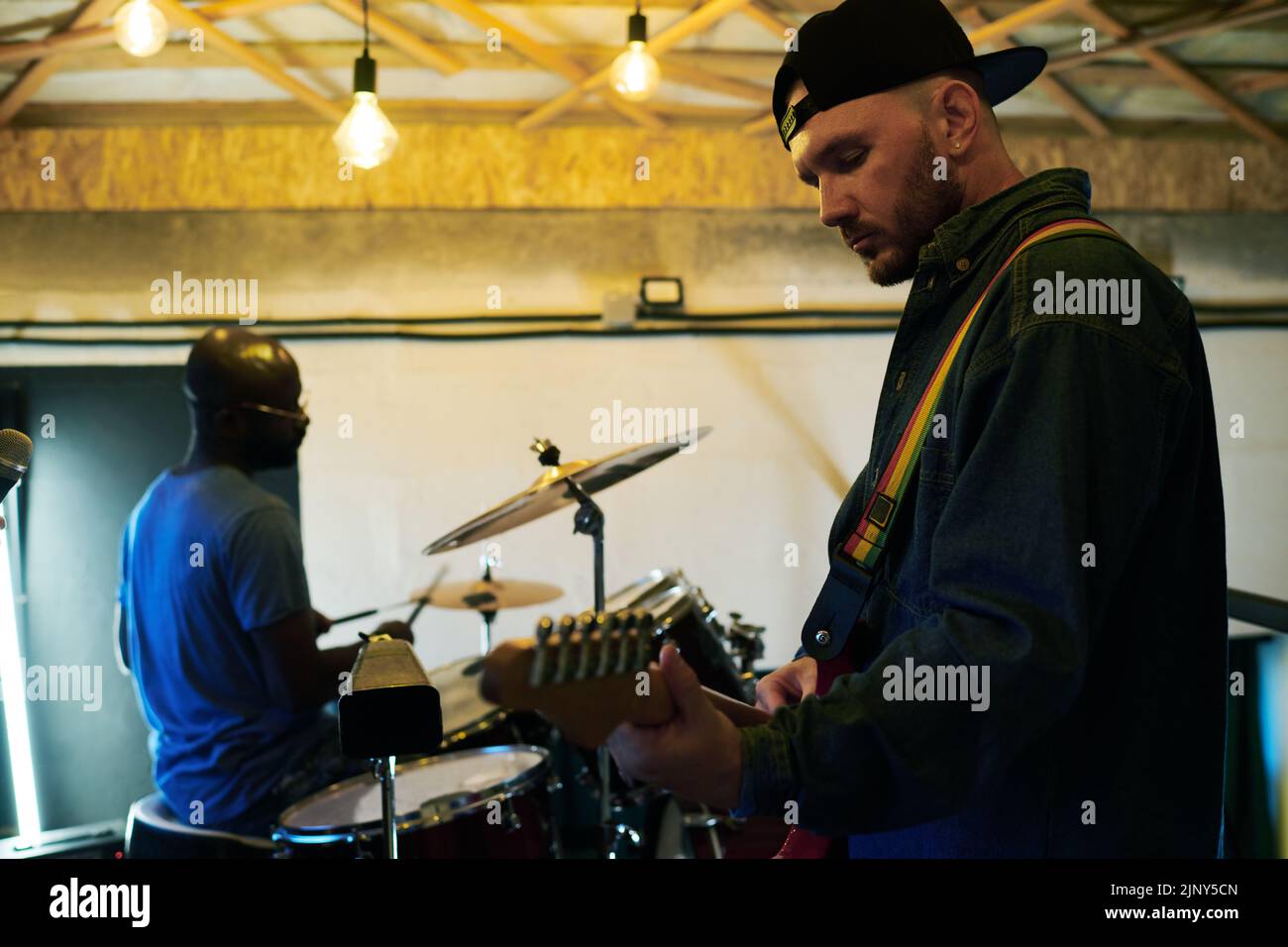 Young man playing electric guitar while standing in front of camera against African American guy hitting cymbals and drums Stock Photo