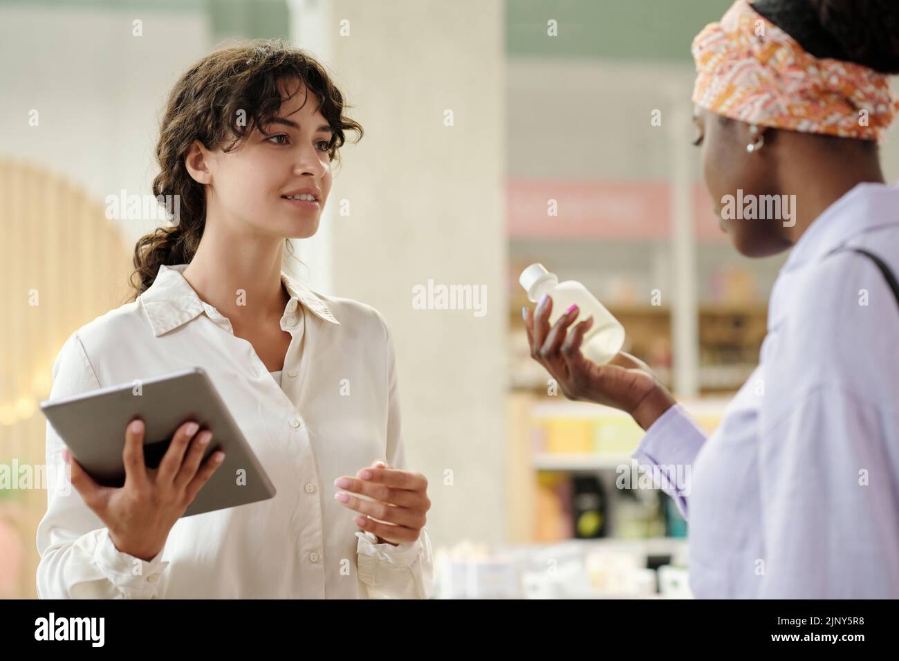 Young saleswoman with tablet looking at female customer with plastic bottle containing liquid self care product and consulting her Stock Photo