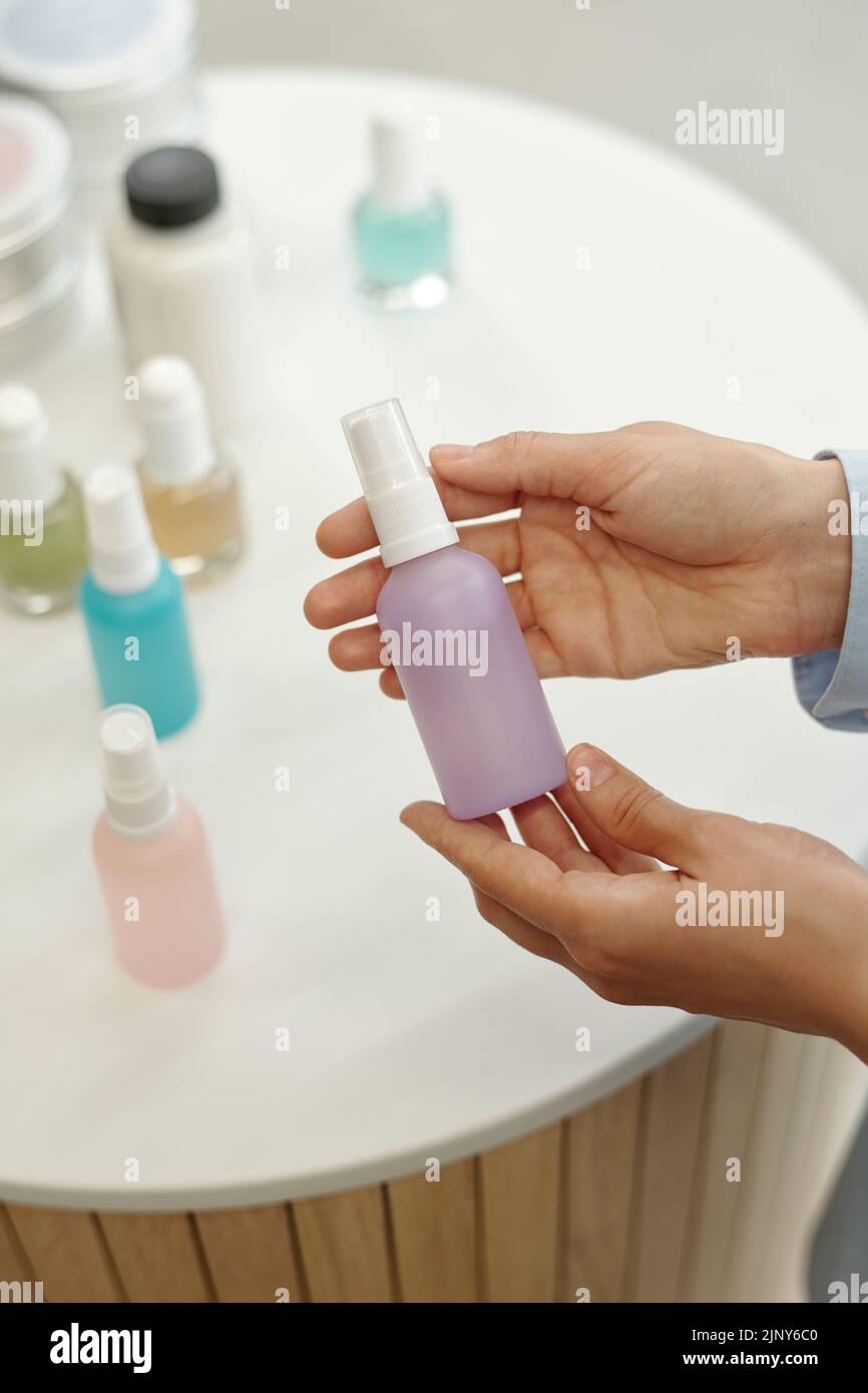 Hands of young woman holding small plastic bottle containing liquid beautycare product of lilac color over display with other items Stock Photo