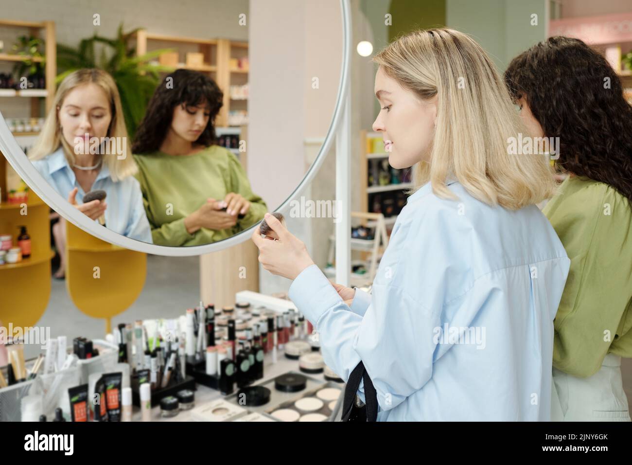 Young blond female shopper holding powder brush while choosing accessories for makeup in front of mirror and display Stock Photo