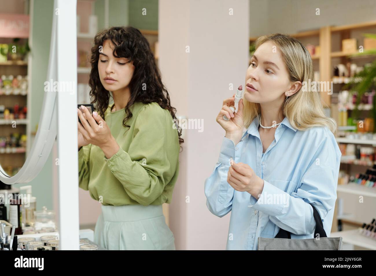 Young blond female consumer testing concealer stick on her face while standing in front of mirror in cosmetic supermarket Stock Photo