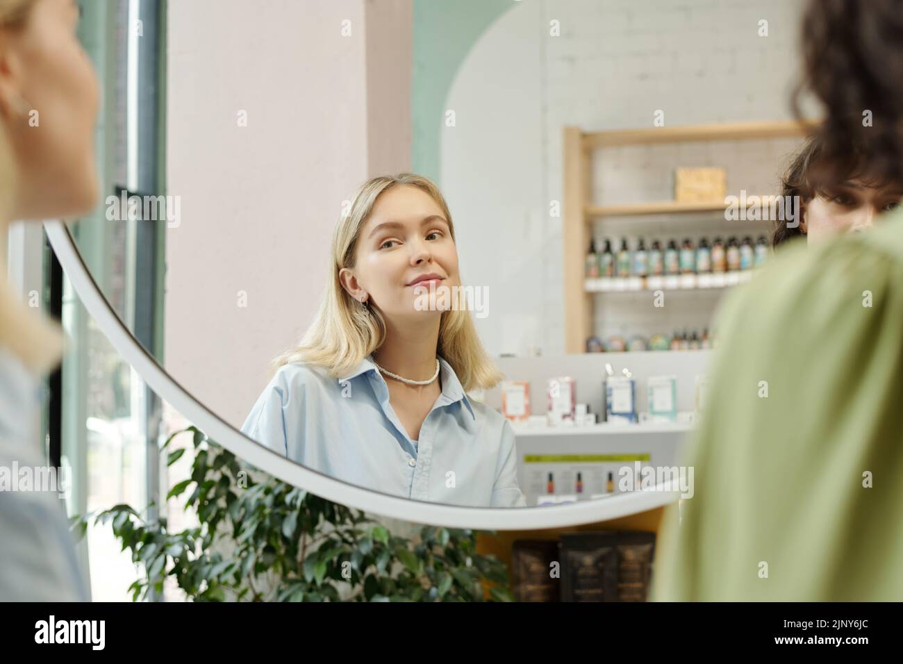 Young pretty woman with long blond hair and natural makeup looking in mirror while testing new beautycare products in cosmetic shop Stock Photo