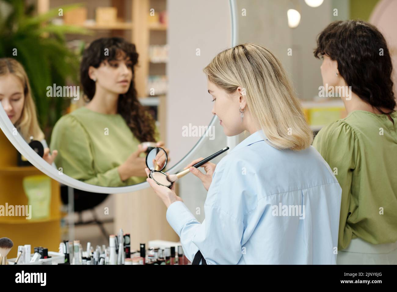 Young customer with brush and compact powder testing beauty products while standing in front of mirror in cosmetic supermarket Stock Photo