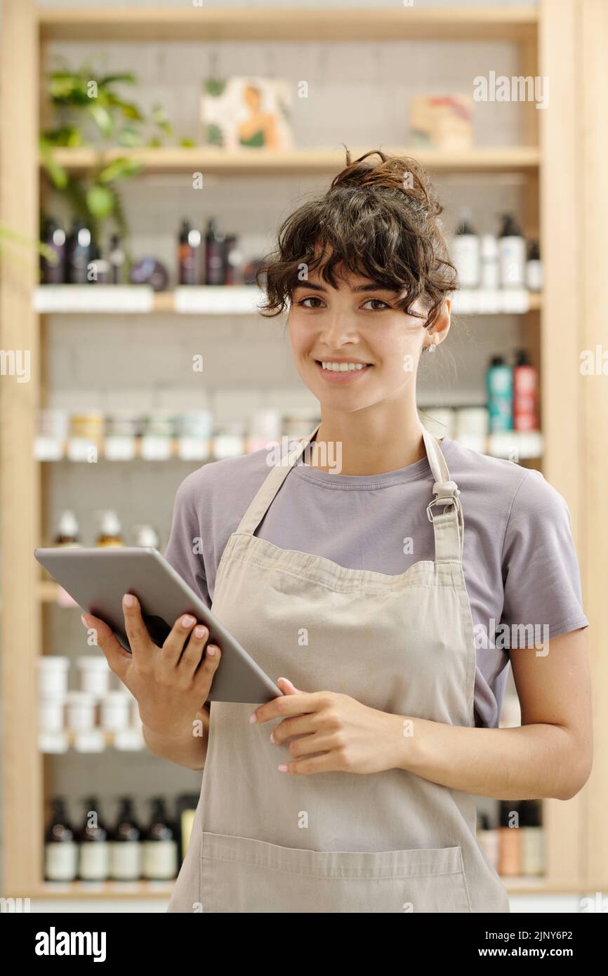 Young successful female shop assistant with digital tablet standing in front of camera against displays with beautycare products Stock Photo