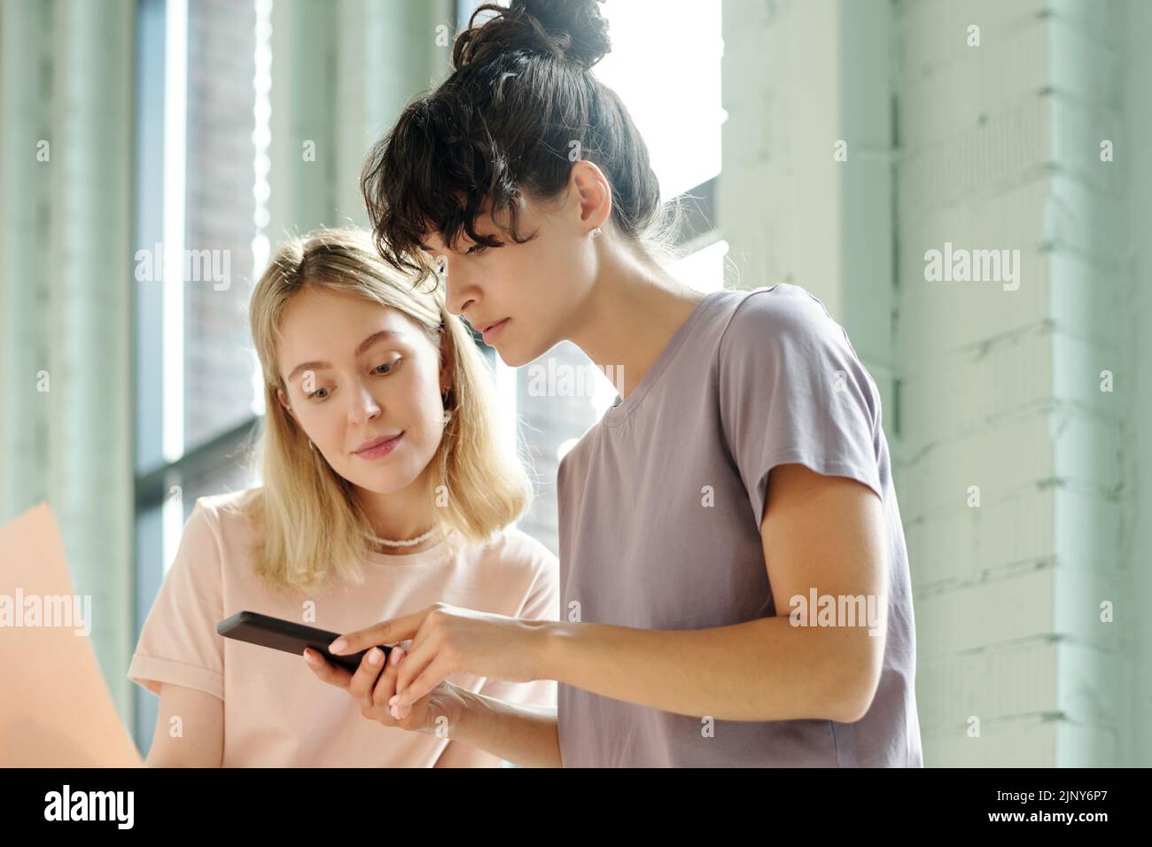 Two young pretty colleagues in t-shirts taking photo of beautycare products on smartphone for advertisement or presentation Stock Photo