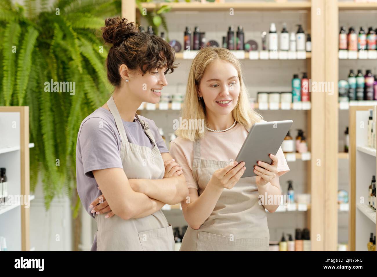Two young smiling shop assistants in workwear looking at tablet screen while discussing assortment or consulting clients online Stock Photo