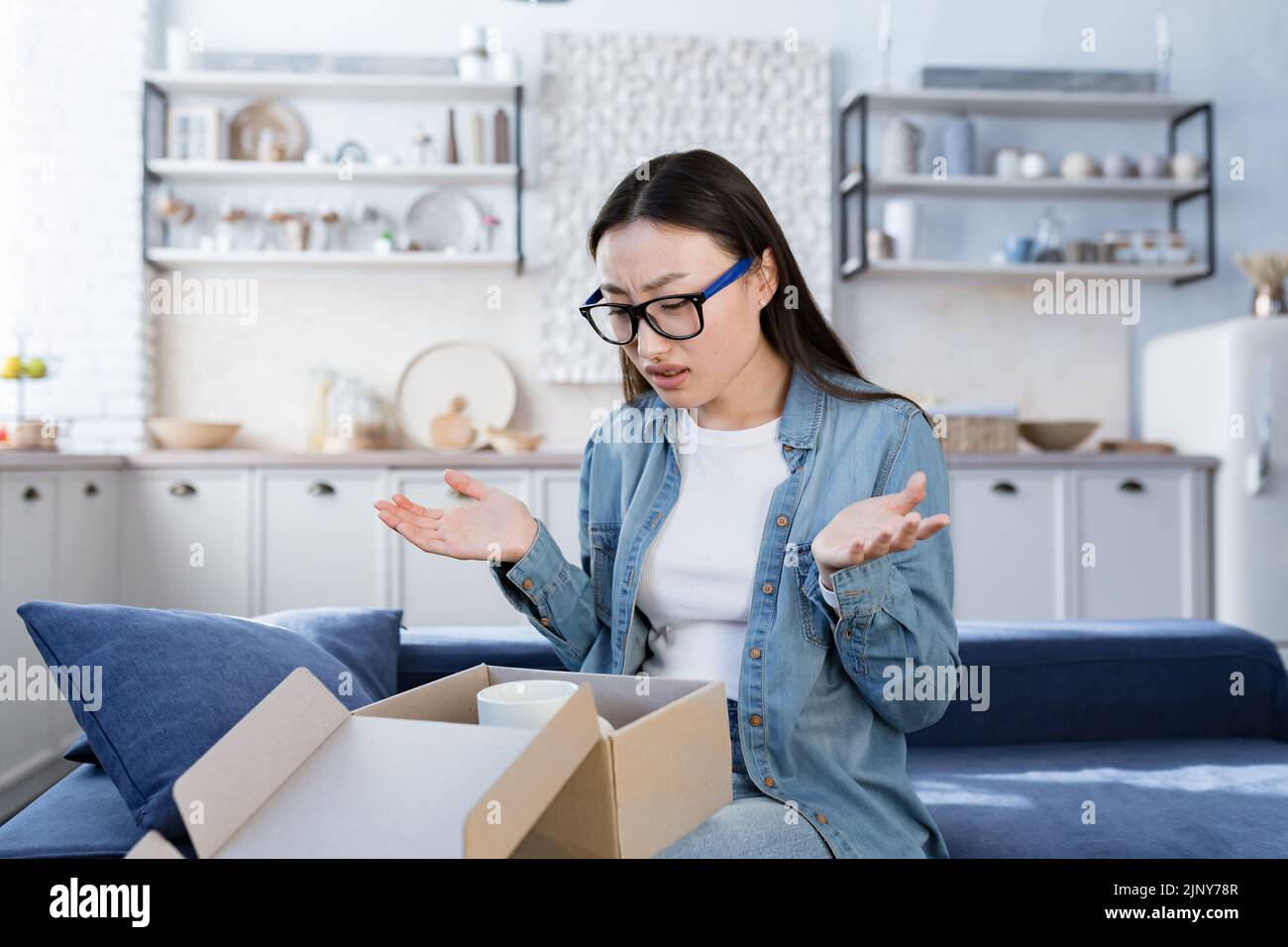 Disappointed woman buyer at home, Asian woman received wrong parcel and damaged product from online store, teenage girl in glasses upset sitting on sofa Stock Photo