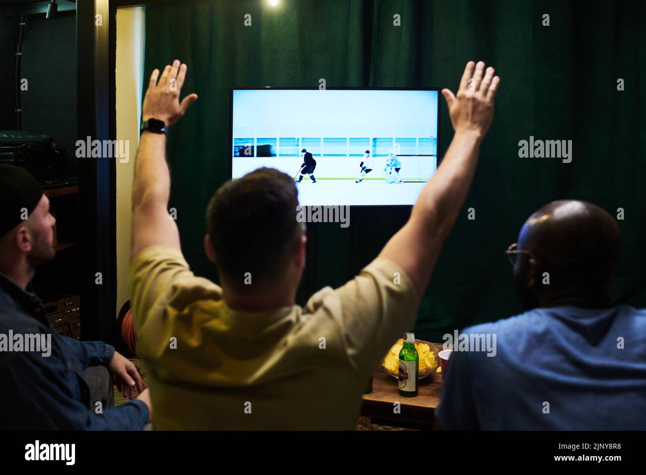 Rear view of three young intercultural men watching hockey broadcast on tv and having beer with snack while one of them keeping arms raised Stock Photo