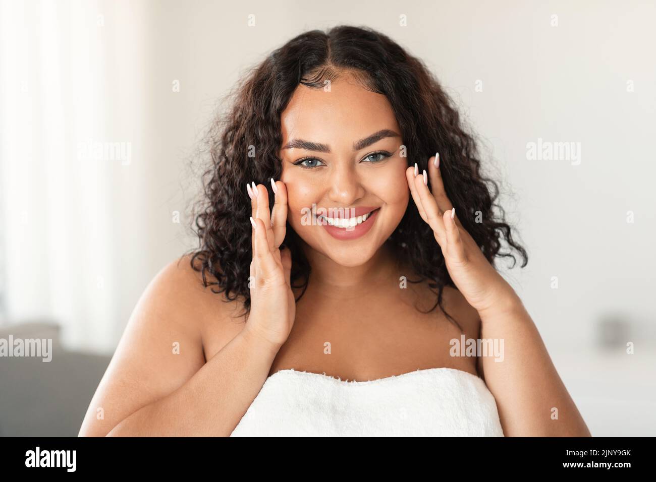 Face massage. African american plus size woman touching her smooth flawless skin on cheeks and smiling at camera Stock Photo