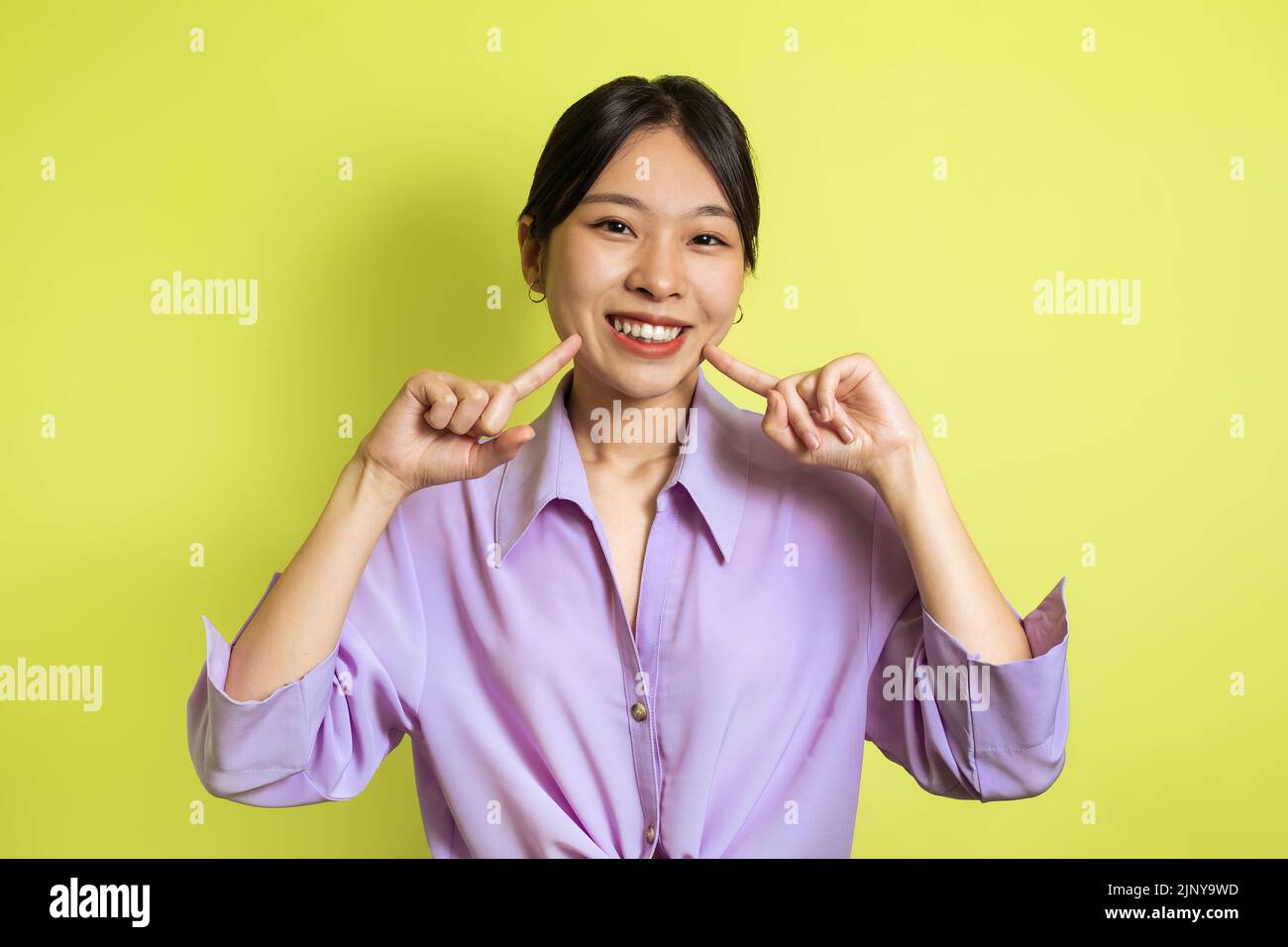 Toothy Asian Female Pointing Fingers At Her Smile, Yellow Background Stock Photo