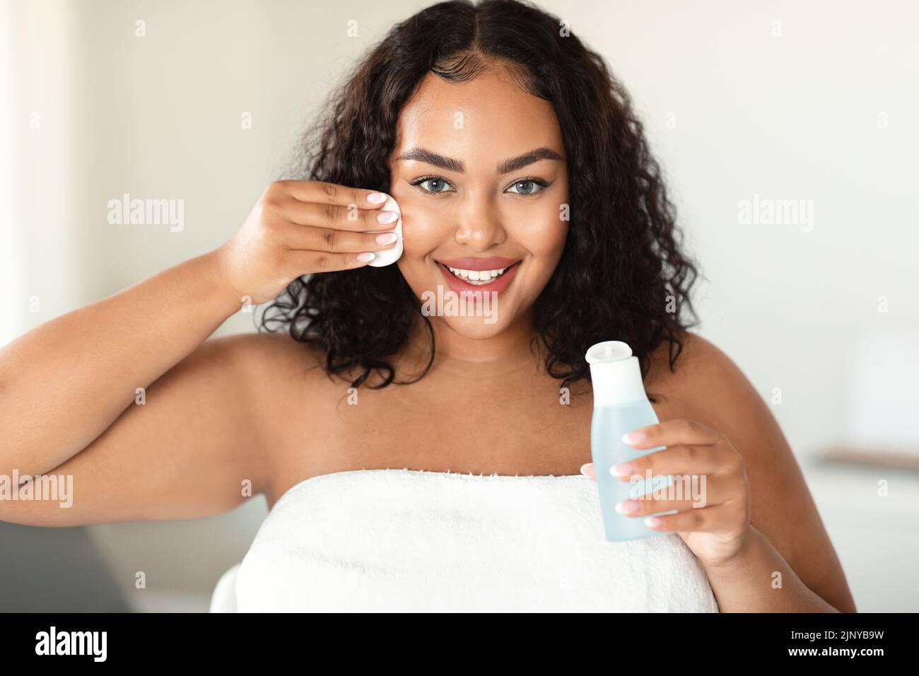 Skin cleansing concept. Happy black bodypositive lady using micellar water and cotton pad, making daily skincare routine Stock Photo