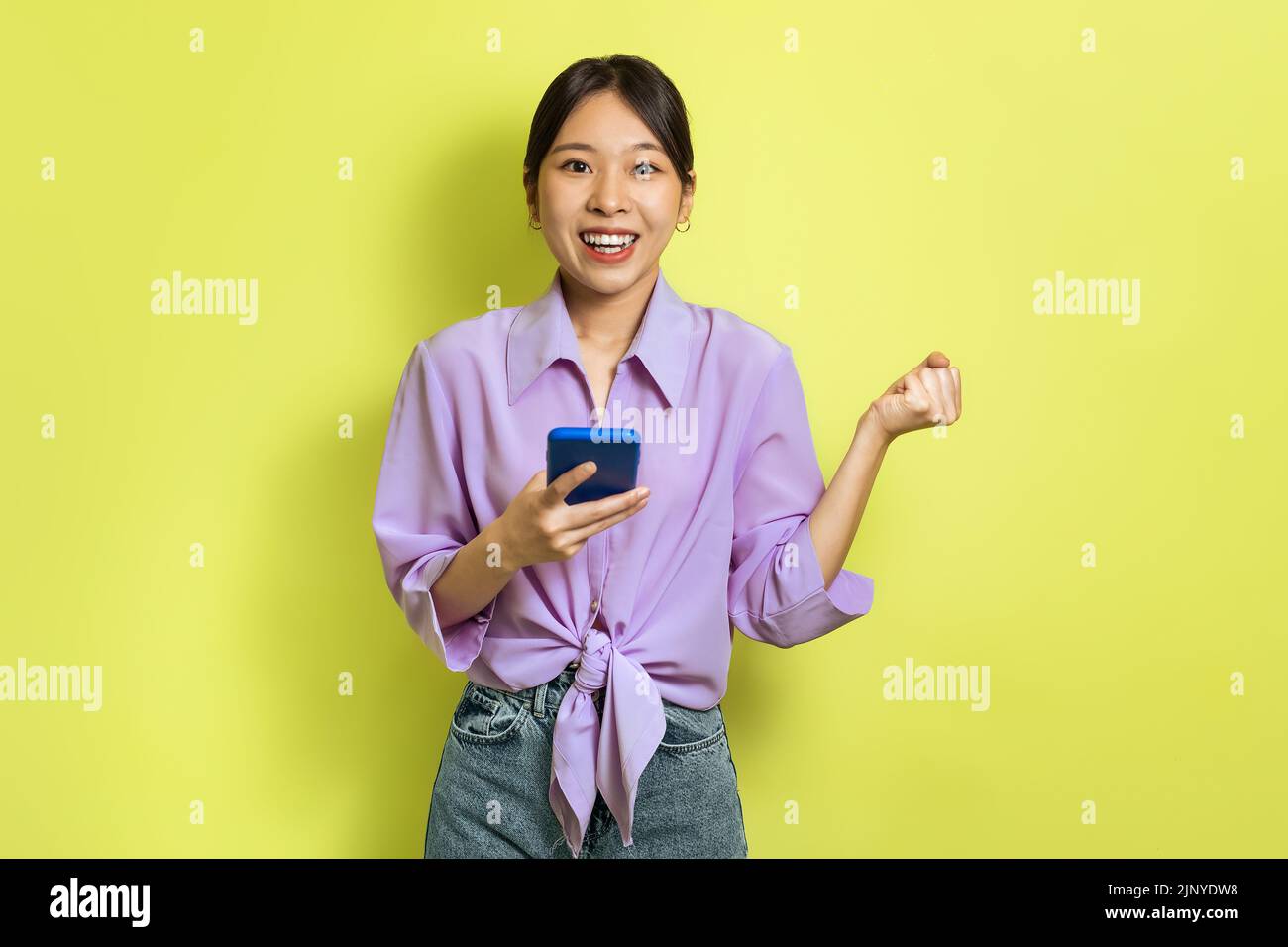 Joyful Asian Lady Using Cellphone And Shaking Fists, Yellow Background Stock Photo