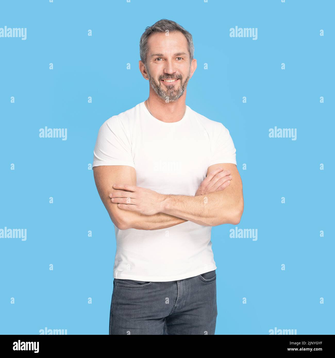 Handsome middle aged grey haired man standing with arms folded happy looking at camera wearing white t-shirt and jeans isolated on blue background. Mature fit man, healthy lifestyle concept. Stock Photo