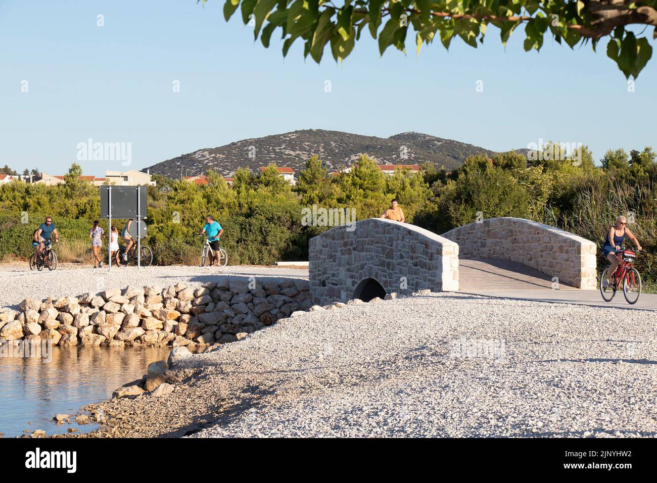 Vodice, Croatia - July 19, 2022: People riding bikes and walking on a coast walkway with a bridge Stock Photo