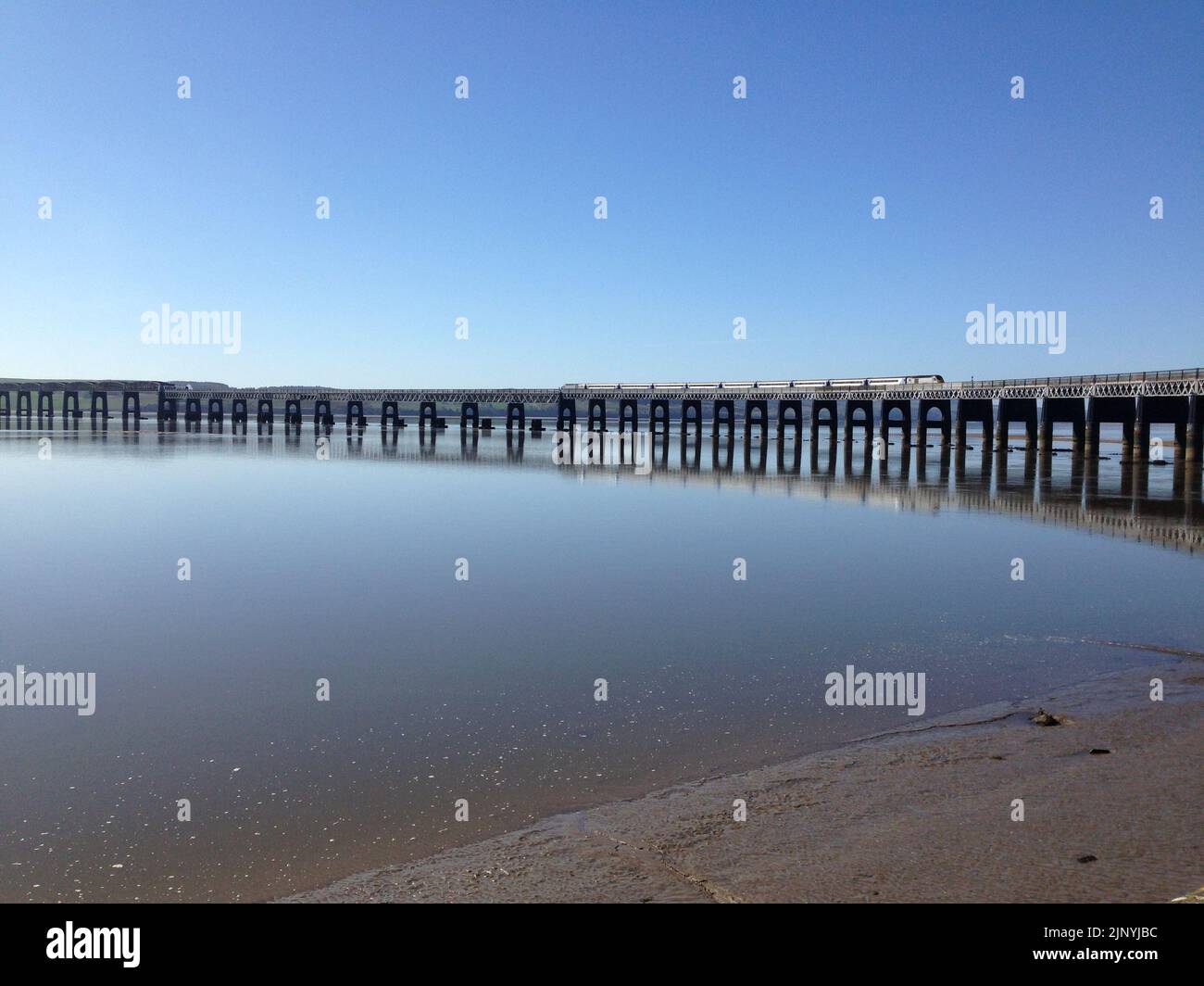 A train on the East Coast Mainline crossing the Tay Bridge, with reflection in the water in calm conditions under blue sky at Dundee, Scotland, UK. Stock Photo