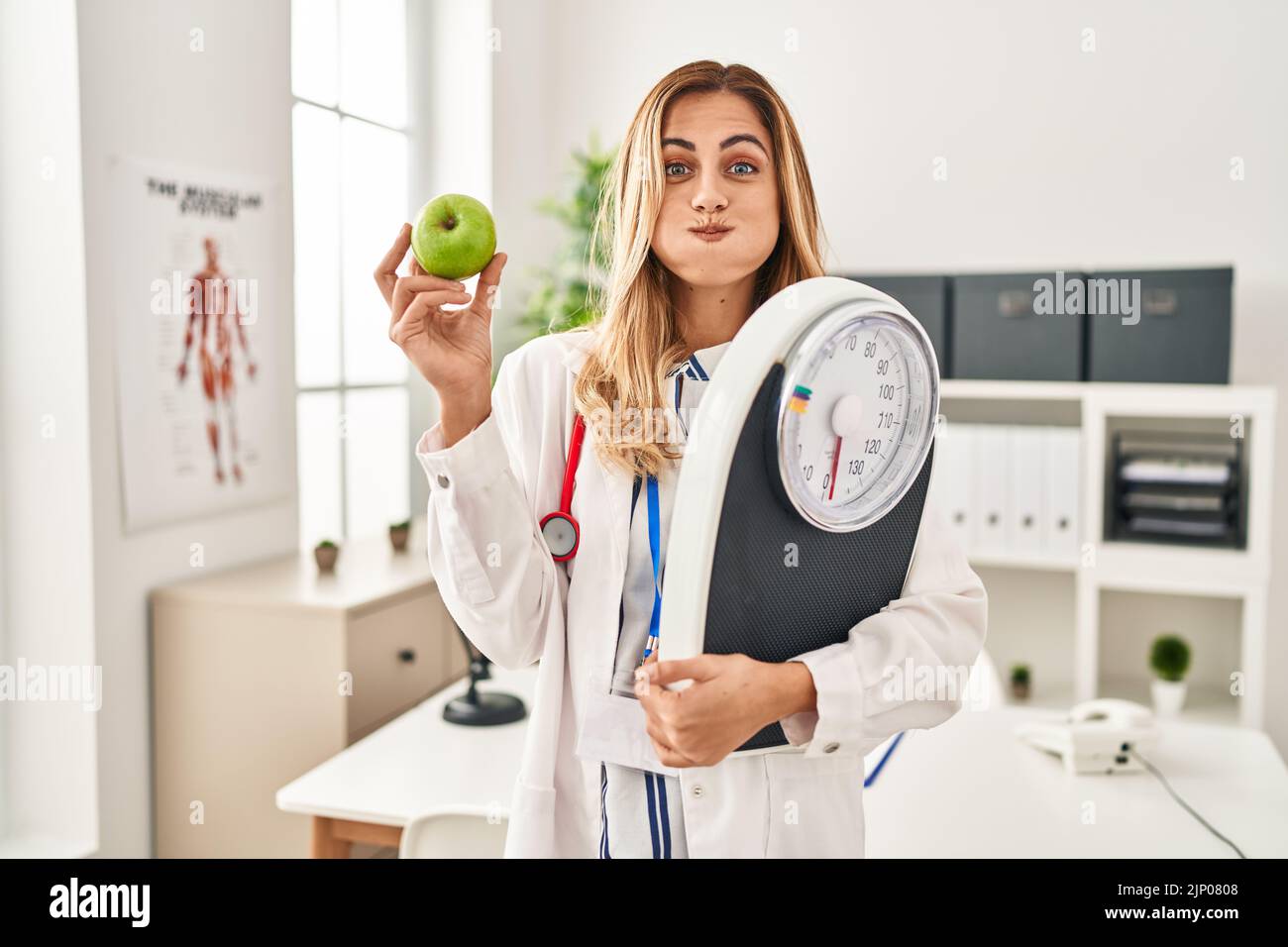 Young blonde doctor woman holding weighing machine and green apple puffing cheeks with funny face. mouth inflated with air, catching air. Stock Photo