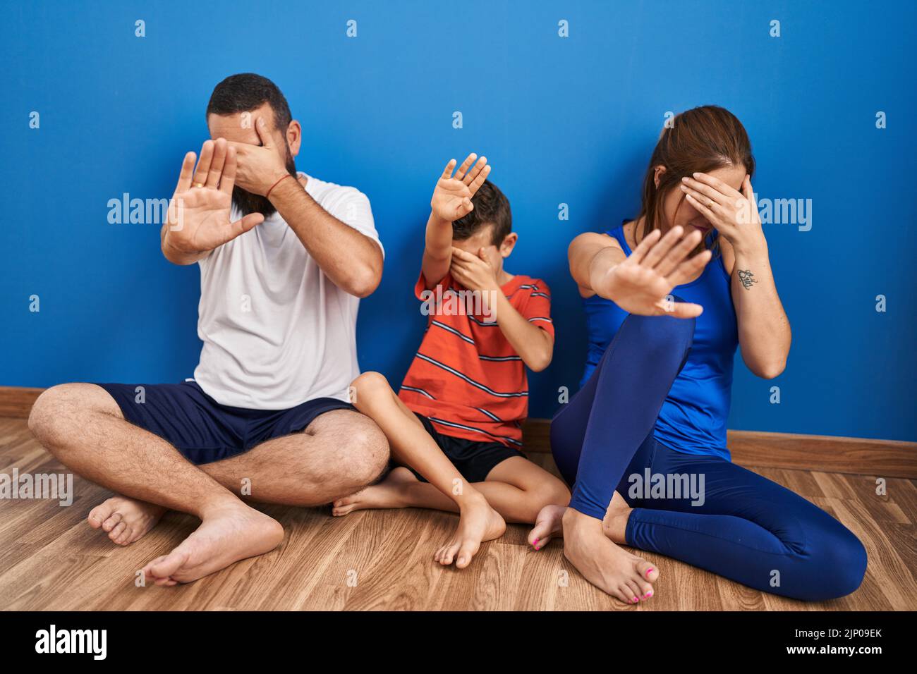 Family of three sitting on the floor at home covering eyes with hands