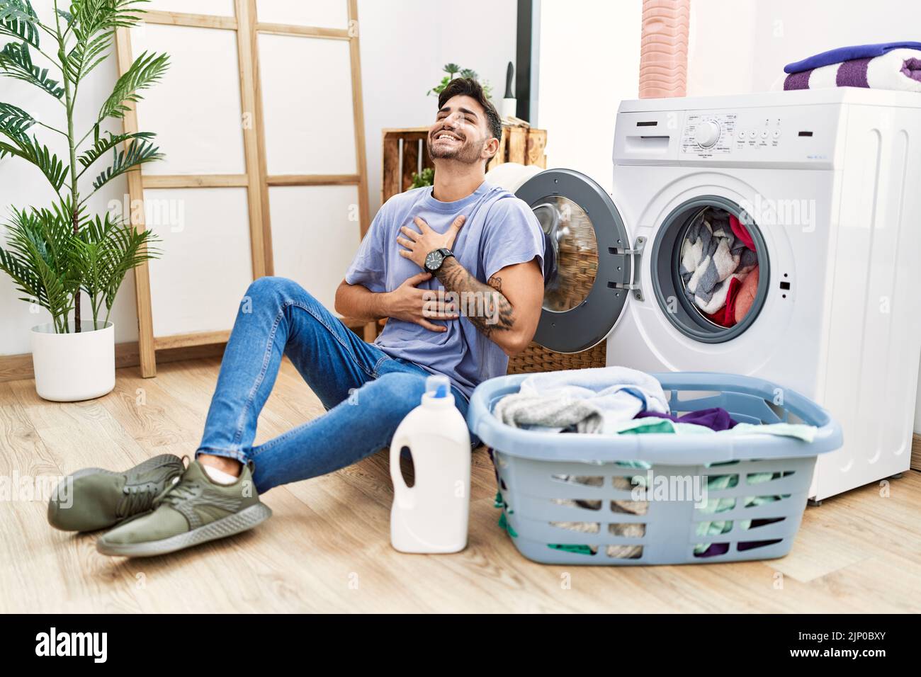 Young hispanic man putting dirty laundry into washing machine smiling